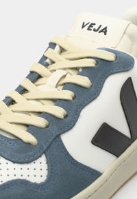 Sapatilhas de camurça azul e couro branco com atacadores em tom creme, acabamento texturizado e um logo preto na lateral. Apresenta detalhes perfurados.