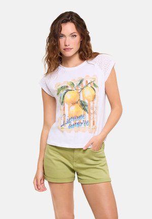 Mujer con cabello ondulado, con una camiseta blanca con estampado de limón y pantalones cortos verdes, de pie con una mano en el bolsillo frente a un fondo liso.