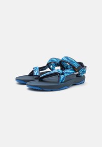 Sandaler med en mørk blå såle og stropper med et blått abstrakt mønster. Materialet virker slitesterkt med en teksturert overflate for bedre grep.