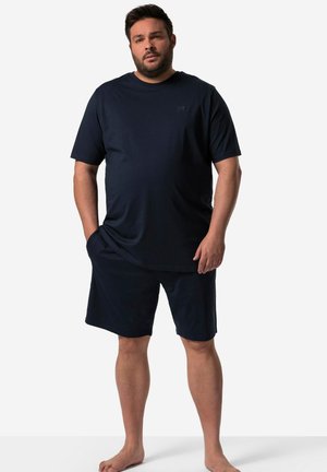 Mann steht barfuß mit den Händen in den Taschen, trägt ein dunkelblaues kurzärmeliges T-Shirt und passende Shorts vor einem einfarbigen hellen Hintergrund.