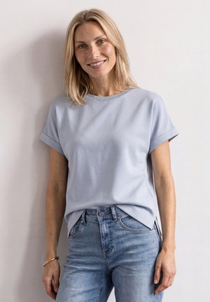 Femme souriante aux cheveux blonds mi-longs, portant un t-shirt bleu clair à manches courtes et un jean bleu, debout devant un mur uni.