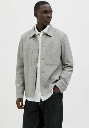 Veste grise à boutons, en tissu texturé, avec deux poches avant et des boutons en bois, portée sur une chemise blanche et un jean noir.