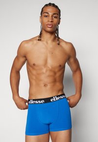 Blaue Boxershorts mit einem schwarzen Bund, der mit weißem "ellesse"-Branding versehen ist. Der Stoff wirkt glatt und passend zum Körper.