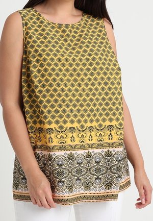 Tunique jaune sans manches avec des motifs floraux et géométriques noirs et blancs complexes, portée avec un pantalon blanc.