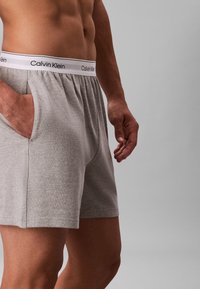 Mandlig overkrop iført grå Calvin Klein boxershorts med elastisk talje, den ene hånd i lommen, stående mod en ensfarvet grå baggrund.