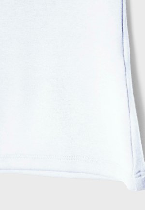 Detailní záběr rohu bílého látkového materiálu s viditelným prošitím a přeloženým okrajem na jednobarevném pozadí.