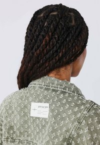 Groene getextureerde jas met een stippenpatroon en een rechthoekig wit etiket op de achterkant met de tekst "PXP." Haar gestyled in nette draaiingen.