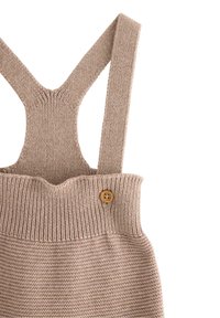 Strikket beige overall med brede stropper, ribbet tekstur ved taljen og en trækknapdetalje på siden.