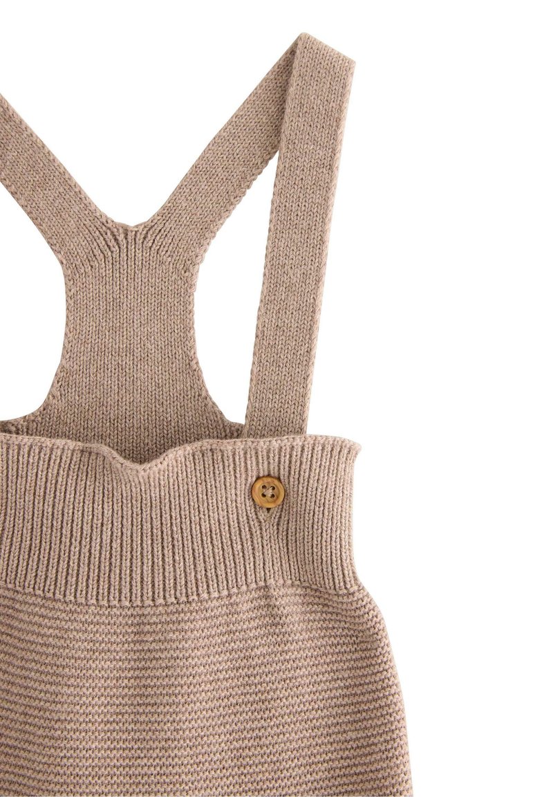 Strikket beige overall med brede stropper, ribbet tekstur ved taljen og en trækknapdetalje på siden.