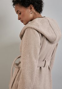 Manteau en mélange de laine beige avec une finition texturée, doté d'une grande capuche et d'une ceinture à la taille. La vue de côté montre la structure et le détail des poches.