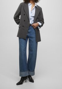 Blazer gris à rayures fines croisé, chemise rayée bleu clair, jean bleu à jambes larges avec ourlets retournés, et bottes noires à talons aiguilles pointues.