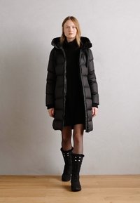 Abrigo largo acolchado negro con capucha, cierre de cremallera en el frente y puños acanalados, llevado sobre un vestido negro y medias, combinado con botas negras hasta la rodilla.