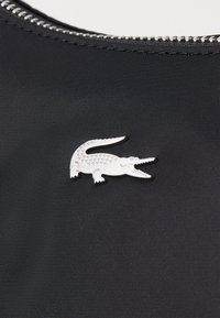 Must värv, millel on sile tekstuur, esitleb hõbedast krokodilli logo pinni, millel on detailid laigud ja avatud suu, kinnitatud metallfekti luku lähedale.
