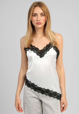Femme aux longs cheveux blonds porte un caraco en satin blanc avec bordure en dentelle noire et un jean gris clair, debout devant un fond gris uni.