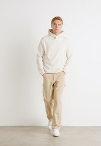 Sudadera con capucha color beige claro de textura acanalada, combinada con pantalones beige y zapatillas blancas. La modelo está de pie contra un fondo liso.