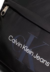 Čierna textilná taška s logom "Calvin Klein Jeans" v bielej a tmavosivej farbe. Obsahuje strieborný zips s akcentom CK.