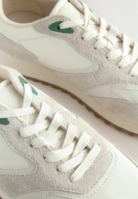 Sneaker mit einem glatten Obermaterial aus weißem Leder und hellgrauen Wildlederelementen. Sie verfügen über grüne Ösen, flache weiße Schnürsenkel und eine strukturierte Gummisohle.