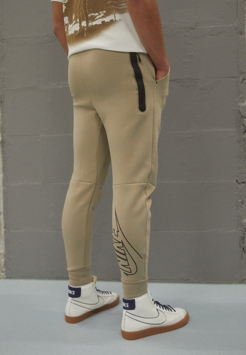 Beige joggingbyxor med slim fit, med en svart sidoknäppning och en stor, svart Nike-logotyp på vänster ben. Matchas med vita högankel-sneakers.
