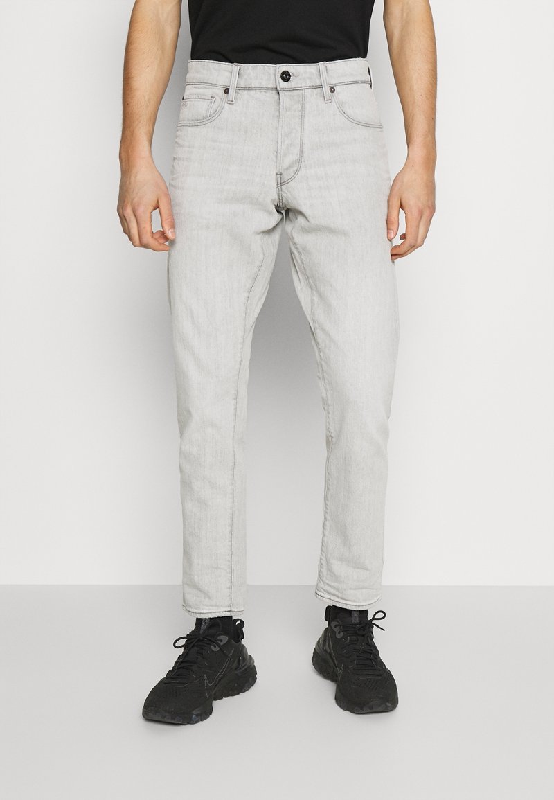 Homme portant un jean slim gris clair et des baskets noires, debout devant un fond blanc uni.
