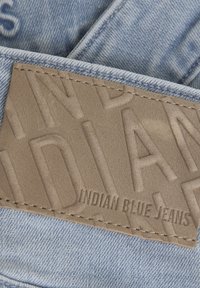 Pezzo in pelle beige con testo in rilievo "INDIAN BLUE JEANS" cucito su denim azzurro chiaro, che mostra una texture liscia e sottili dettagli di cucitura.