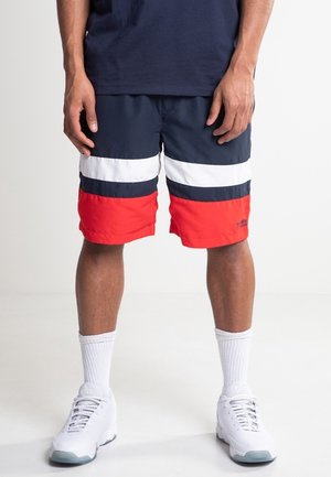 Hombre vistiendo pantalones cortos en bloques de color azul marino, blanco y rojo, calcetines deportivos blancos y zapatillas blancas, de pie sobre un fondo liso.