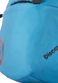 Zaino blu con una texture e forma arrotondata. Presenta un piccolo logo e una finitura liscia. Le cinghie regolabili sono attaccate nella parte superiore.