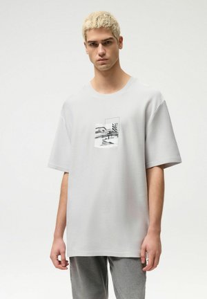 Młody mężczyzna o krótkich blond włosach, ubrany w luźną jasnoszarą koszulkę z czarno-białym motywem architektonicznym z przodu, stojący na białym tle.