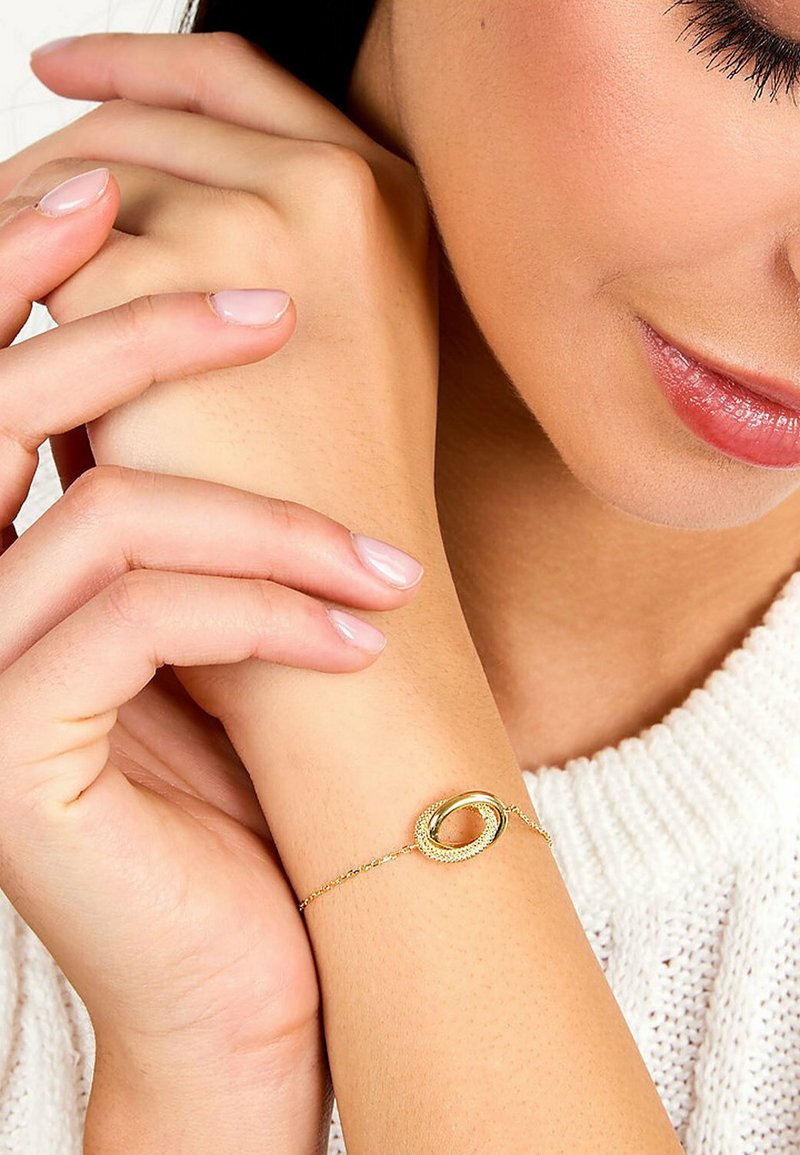 Bracelet en or avec un médaillon ovale texturé et une chaîne délicate, porté sur un poignet à la peau claire, sur un tissu tricoté blanc.