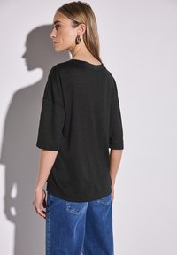 Černé oversized tričko s krátkými rukávy, hladkou texturou, volným střihem a bočními rozparky, doplněné modrými džínovými kalhotami.