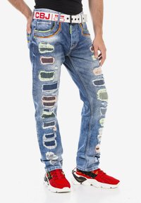 Jeans aus Denim mit einer hellblauen Waschung, die bunte Patchwork-Akzente, distressed Details und einen kontrastierenden weißen Gürtel mit rotem Branding aufweisen.