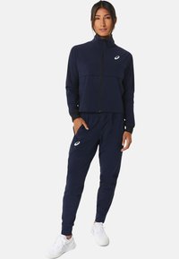 Marineblaue Sportanzughose aus flexiblem Stoff; Jacke mit Reißverschluss und hohem Kragen sowie Hose mit elastischem Bund und Taschen.