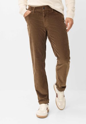 Uomo con pantaloni di velluto marrone, maglione crema e scarpe da ginnastica bianche con dettagli marroni che cammina con una mano in tasca.