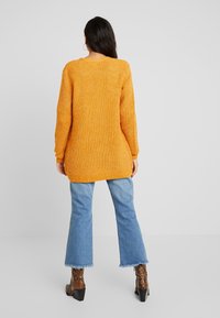 Frau mit langen dunklen Haaren, die einen übergroßen senfgelben Strickpullover, cropped blaue Jeans und braune Schlangenleder-Stiefeletten trägt, stehend von hinten.
