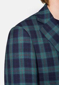 Marynarka w kratę w ciemnoniebieskim i zielonym kolorze z czerwonymi akcentami. Posiada wyraźnie zarysowane ramiona i ostre klapy. Teksturowany tkanina tkana z wzorem w kratę.