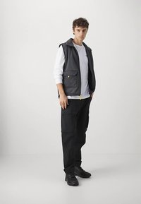 Schwarze Cargo-Hosen, weißes Langarmshirt und eine dunkelgraue gesteppte Weste mit gelbem Reißverschluss, stehend vor einem schlichten Hintergrund.