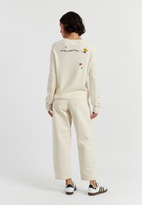 Maglione crema con dettagli ricamati, inclusi un personaggio e cuori, abbinato a pantaloni beige e sneakers bianche a strisce nere.