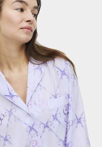 Helle lila Schlafanzugoberteil mit einem Druck aus Schleifen und Wirbeln. Verfügt über einen Reverskragen, eine Knopfleiste auf der Vorderseite und eine kleine aufgesetzte Tasche.