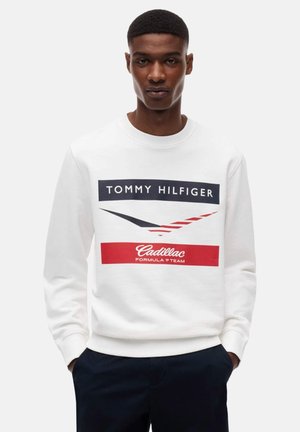 Ung mand iført en hvid sweatshirt med "Tommy Hilfiger" og "Cadillac Formula 1 Team" logoer, hænder i lommerne, neutral baggrund.