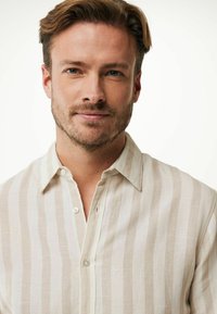 Beige randig linne skjorta med knäppkrage och synliga knappar. Mjuk textur med jämnt fördelade vertikala ränder som skapar ett subtilt mönster.