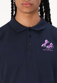 Navy blue polo shirt in cotton, featuring a three-button placket. Pink embroidered unicorns and brand name "CLEPTOMANIX" displayed on the chest.