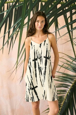 Naine seisab suurte roheliste palmilehtede ees, kandes valget varrukateta kleiti mustade abstraktsete ristmustritega.