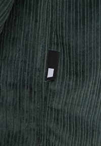 Groene ribstof met een zachte textuur, met een klein zwart label met een witte rechthoek. Close-up opname met focus op het materiaal.