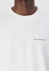 Tricou alb din bumbac cu guler rotund, având un mic logo negru "Abercrombie | NYC" pe partea stângă. Textură netedă.