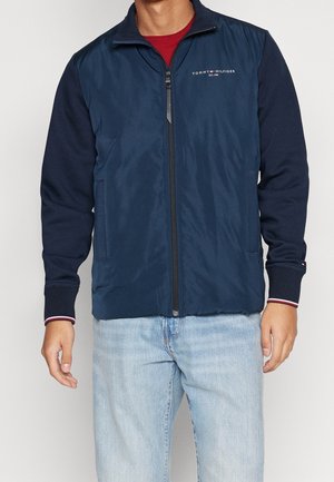 Mand iført navy blå lynlåsvest over mørk langærmet skjorte med Tommy Hilfiger-logo, kombineret med lyseblå jeans.