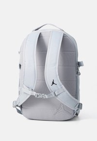 Gray backpack featuring padded compartments, adjustable straps, and a black logo detail. The material has a smooth texture with a subtle pattern.