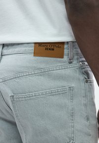 Svetlosivé rifľové nohavice s hnedou koženou nálepkou s nápisom "Marc O'Polo DENIM", obsahujúce zadné vrecko a viditeľné šijacie detaily.