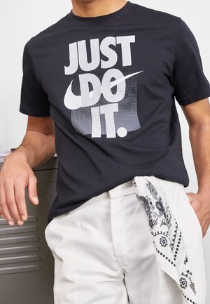 Mężczyzna ubrany w czarną koszulkę Nike z napisem i logo "JUST DO IT.", białe spodnie oraz czarno-białą bandanę włożoną do nich.