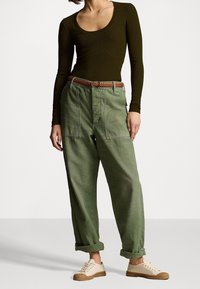 Pantalones cargo de corte suelto en verde oliva, con puños enrollados, que cuentan con grandes bolsillos laterales y un cinturón tejido marrón anudado a la cintura.