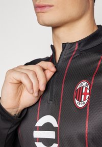 Maglietta sportiva nera con strisce rosse, colletto alto e chiusura a zip. Presenta un logo circolare con design rosso e nero sul petto.