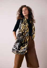 Frau mit lockigem Haar, die ein schwarzes Oberteil, braune, hoch taillierte Shorts und einen gelb-schwarzen Schal mit Tierdruck trägt, seitlich schaut vor einem einfarbigen Hintergrund.